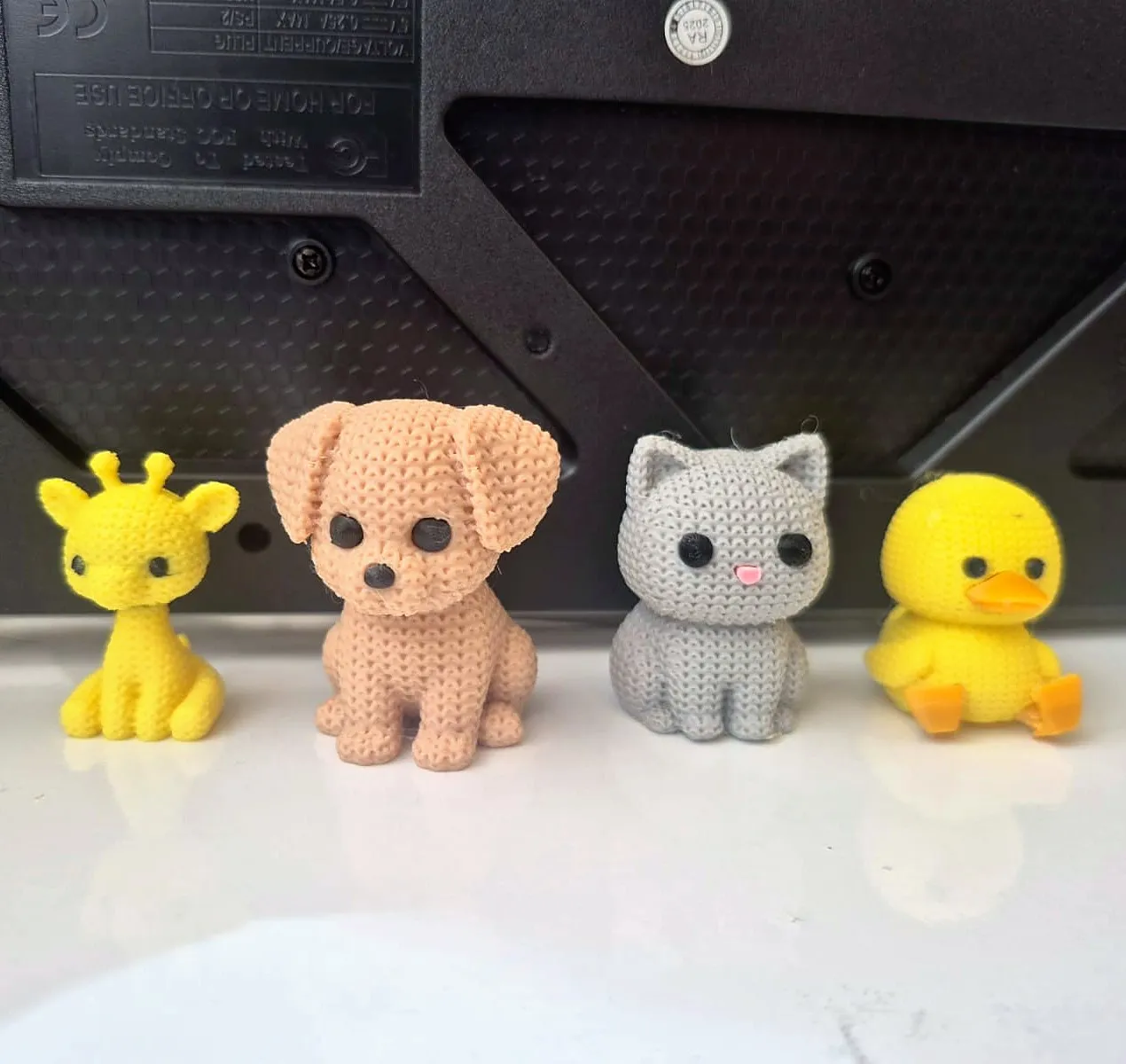 Quatro bichinhos impressos em 3D com textura de tricô, uma girafa amarela, um cachorro bege, um gato cinza e um pato amarelo