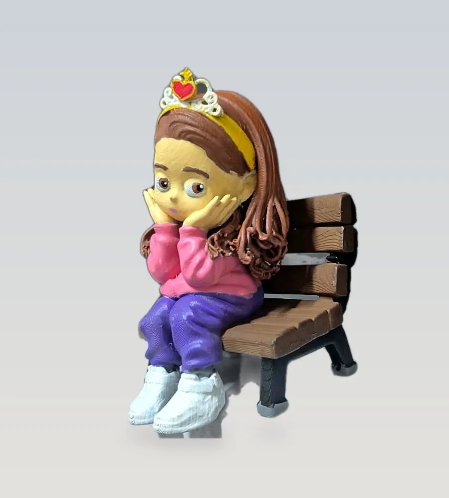 Boneca personalizada de uma menina de cabelo comprido e coroa, sentada em um banco de praça em miniatura