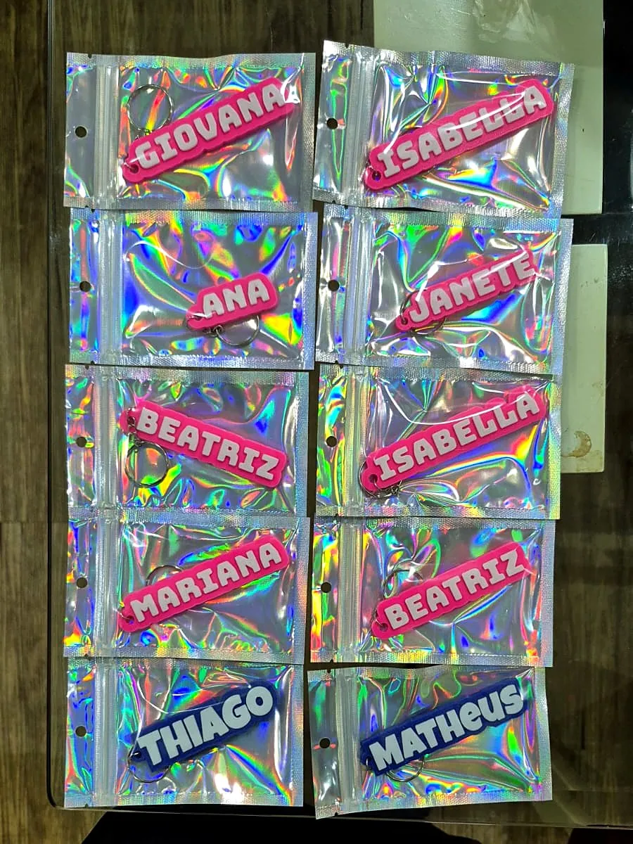 Dez chaveiros com nomes em rosa e azul, cada um embalado em saquinho holográfico