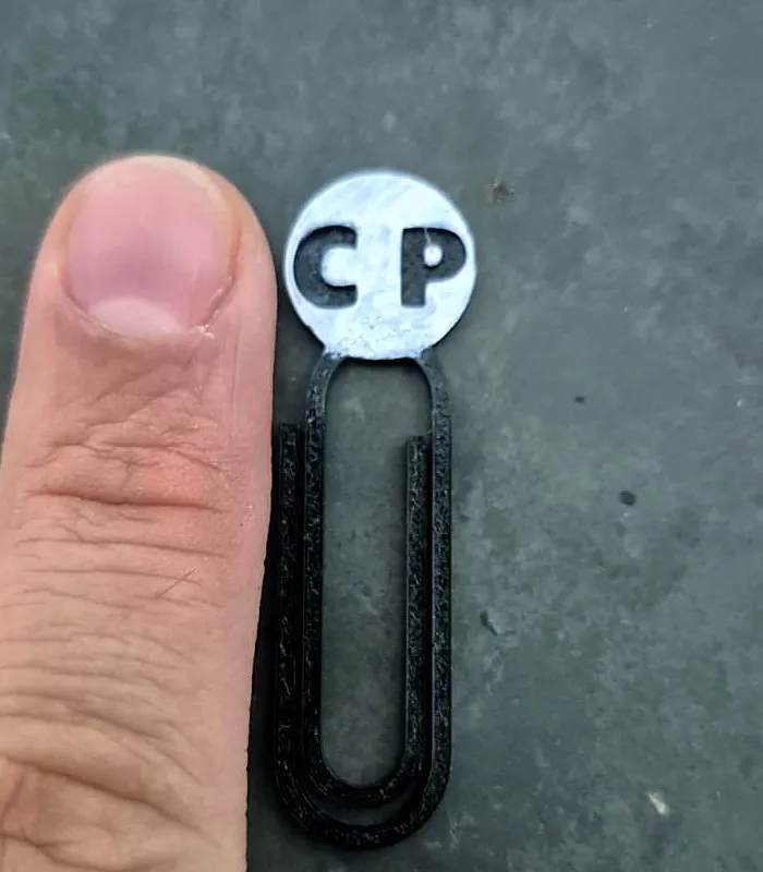 Marcador de página preto em formato de clipe com as iniciais CP em um círculo branco, ao lado de um dedo para mostrar o tamanho