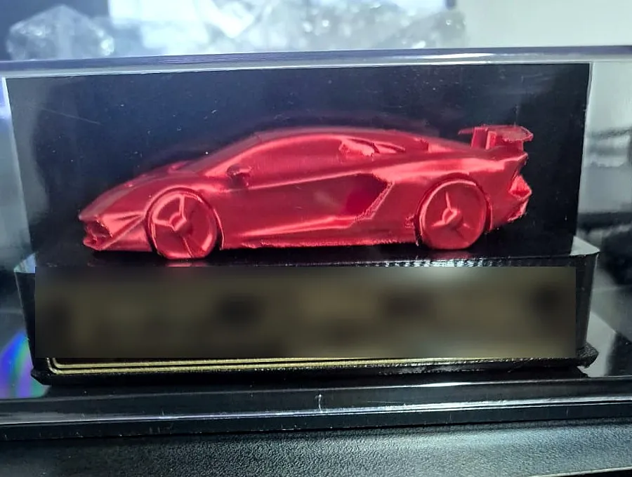 Miniatura de carro esportivo vermelho dentro de caixa de acrílico, sobre base preta com placa dourada (texto da placa desfocado)