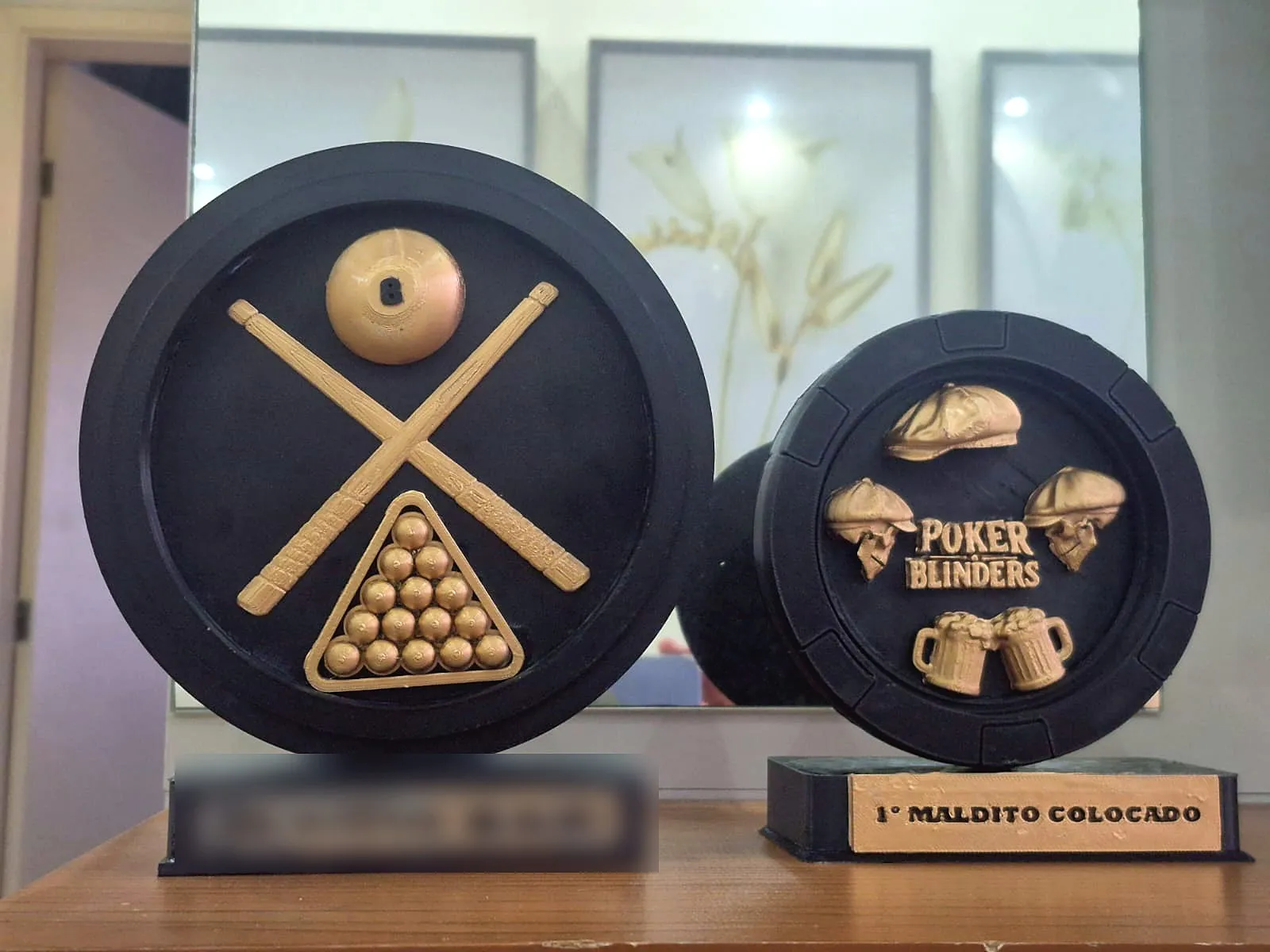 Dois troféus redondos pretos com relevo dourado: um com tacos e bolas de sinuca, outro com o tema Poker Blinders (nome do bar desfocado)